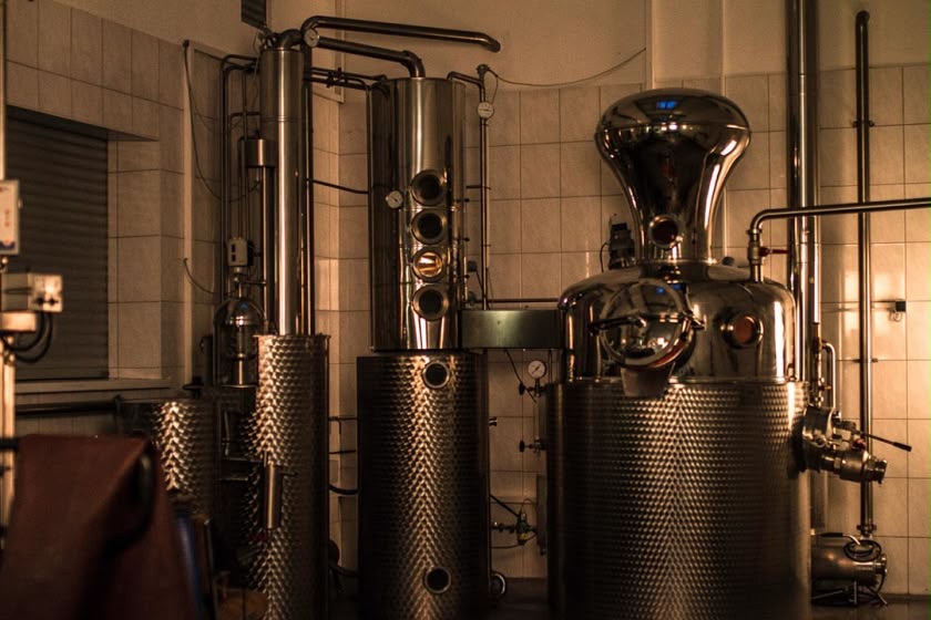 Brennblasen und Edelstahltanks einer kleinen Manufaktur, warm ausgeleuchtet vor weiß gefliesten Wänden.